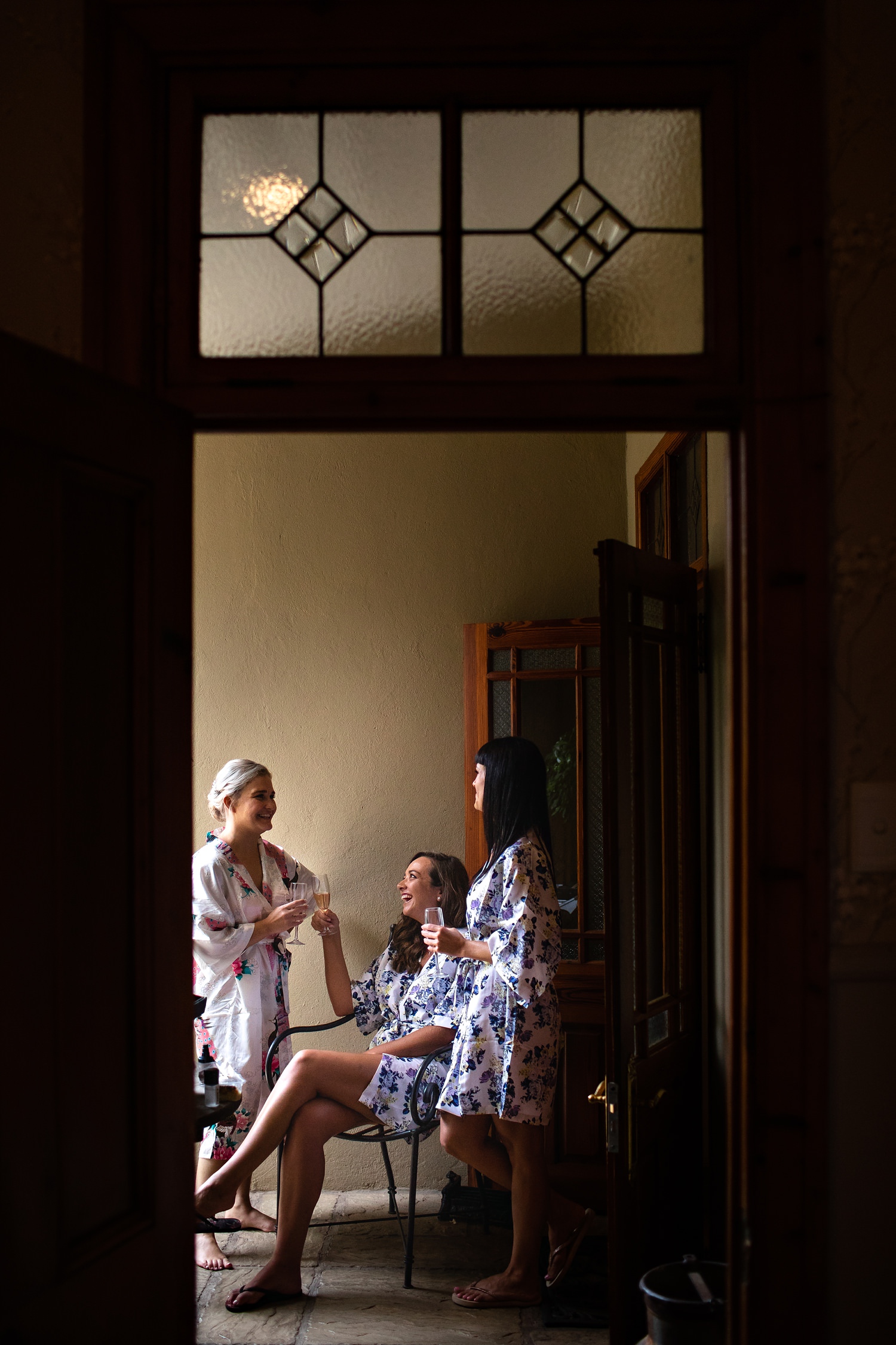 A bride laughs and toasts champagne with her two bridesmaids to celebrate love, dressed in floral silk gowns at Morrells wedding venue, image framed by an antique door