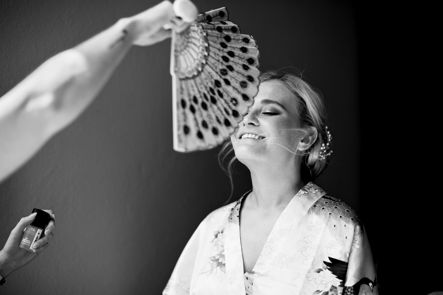 A bride smiles as the make-up artist uses a fan to dry the setting spray applied to her face as the final step by a wedding photographer in South Africa, Niki M