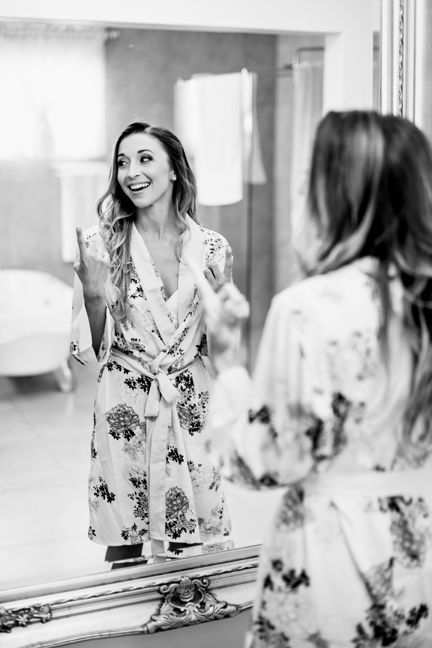 A bride with loosely curled hair stands in front of a mirror and gushes after seeing her natural bridal make-up for the first time since application at Blomfontein Wedding Venue