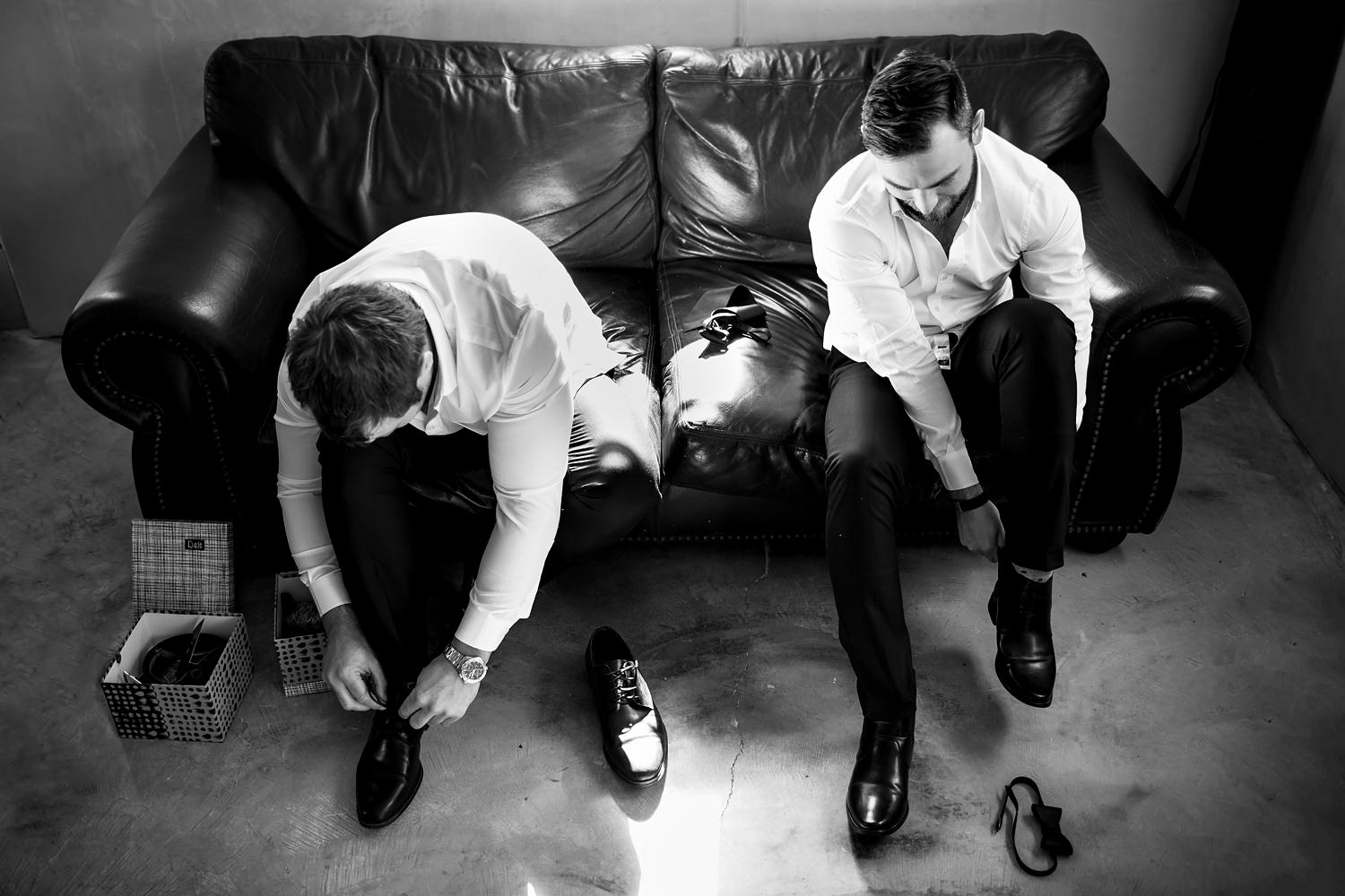 A black and white candid image of the groom and a groomsman sitting on a sofa and putting their shoes on in the light of the window at The Rose Barn
