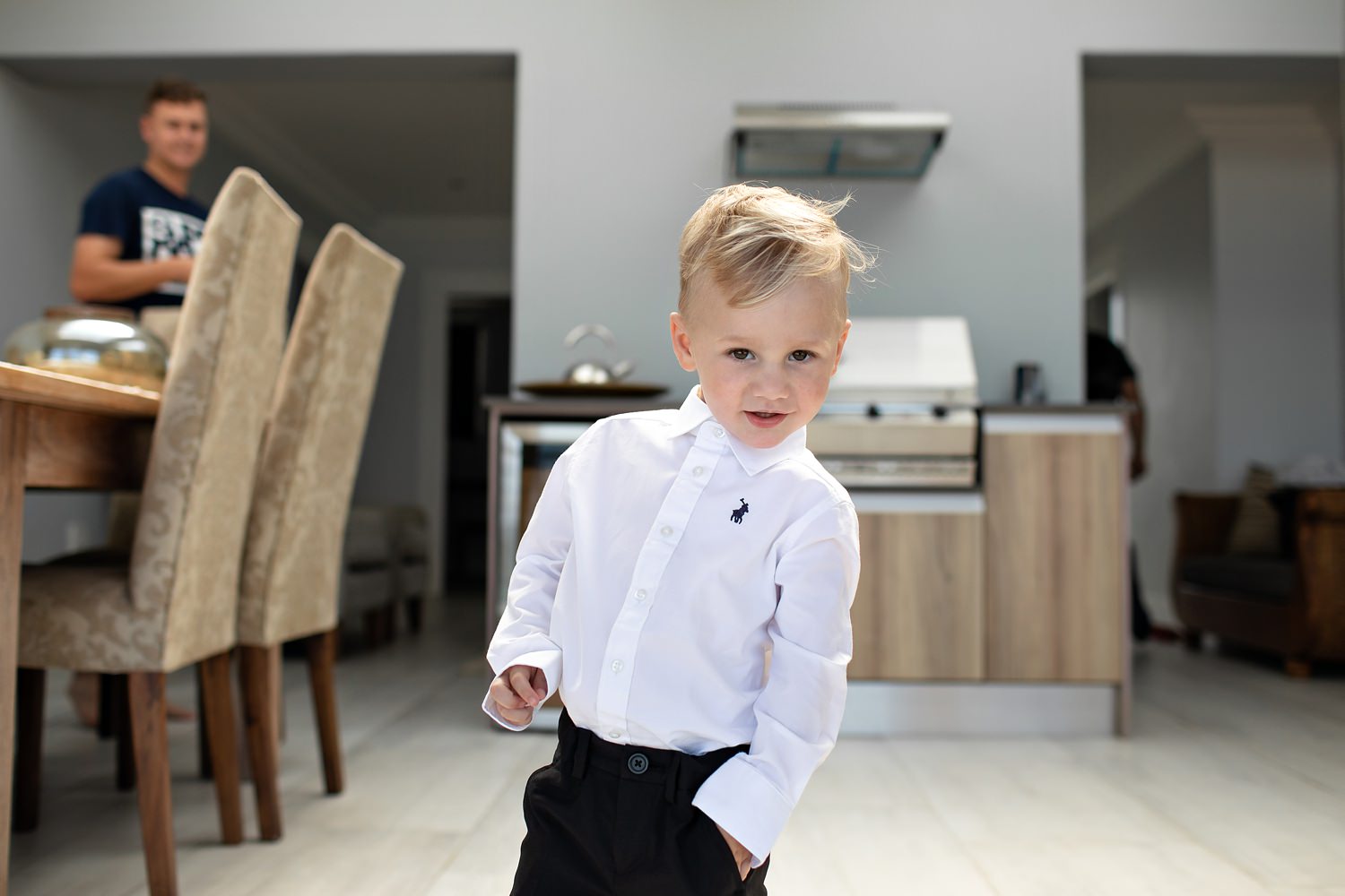 A small ring bearer poses for the camera with one hand in his pocket whilst wearing a white polo shirt