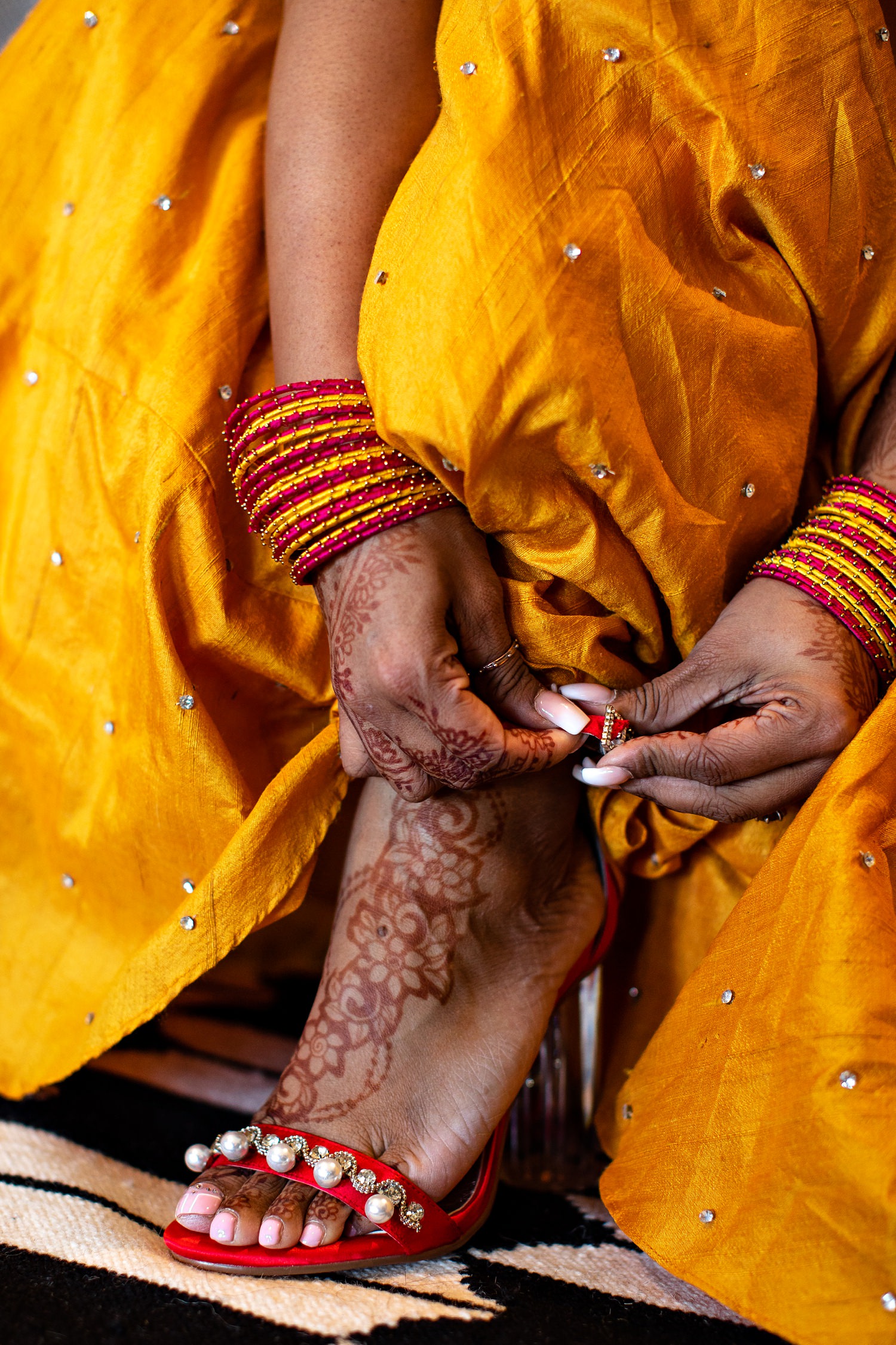 A Hindu Indian bride lifts her mustard yellow dress to expose henna decorations on her feet whilst she fastens the buckle on her red Louboutin shoes