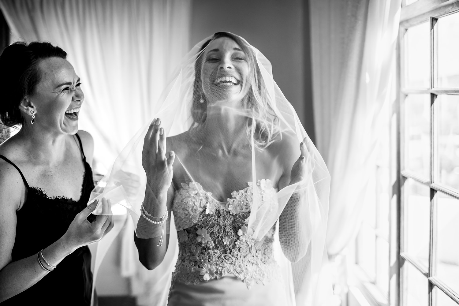 A veiled bride at Blomfontein is overcome with emotion as she lifts her hands to the sky with a big smile and closed eyes, the bridesmaid looking on with joy.