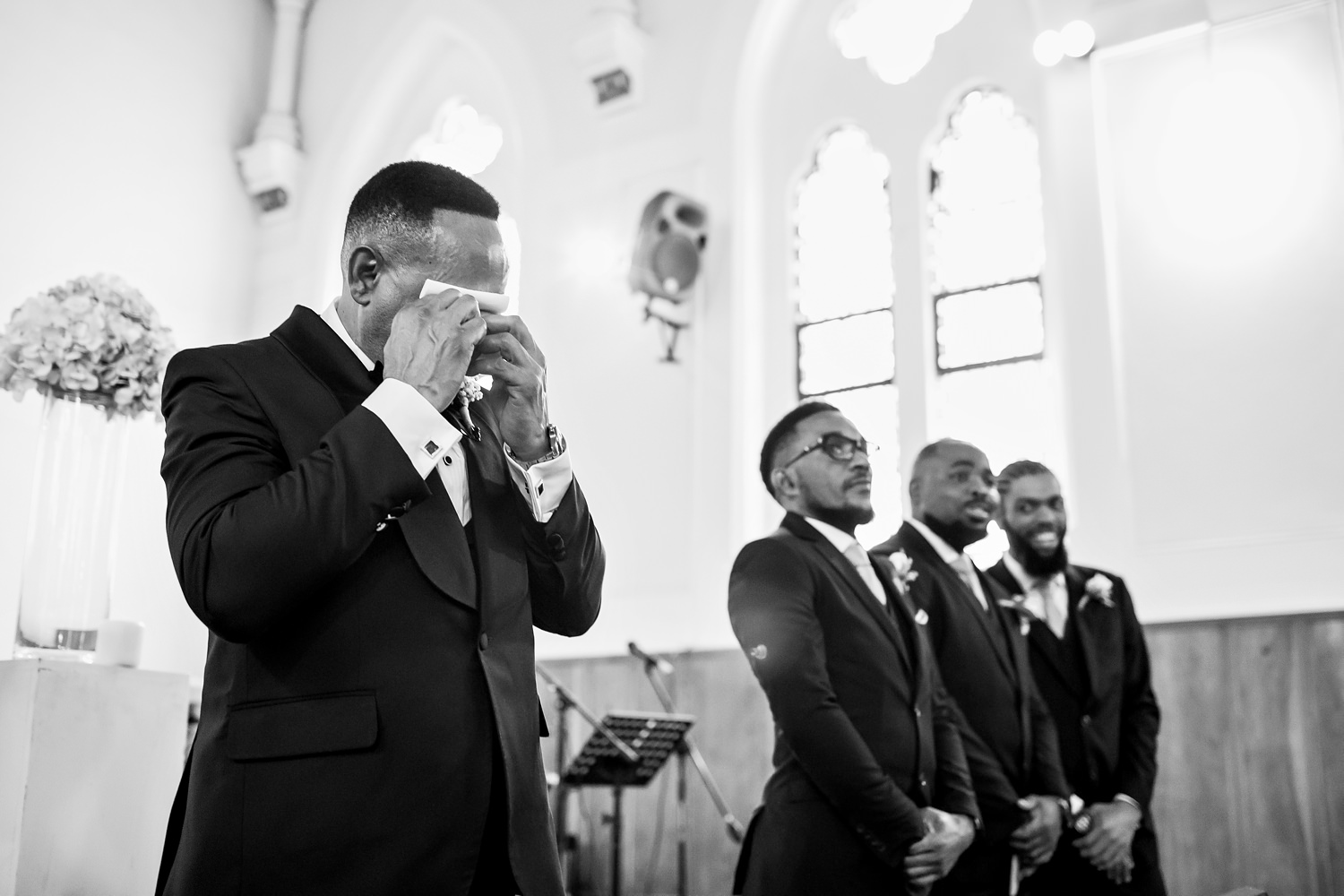 The groom wipes away his tears and gromsmen look on with smiles as the bride makes her way down the church aisle