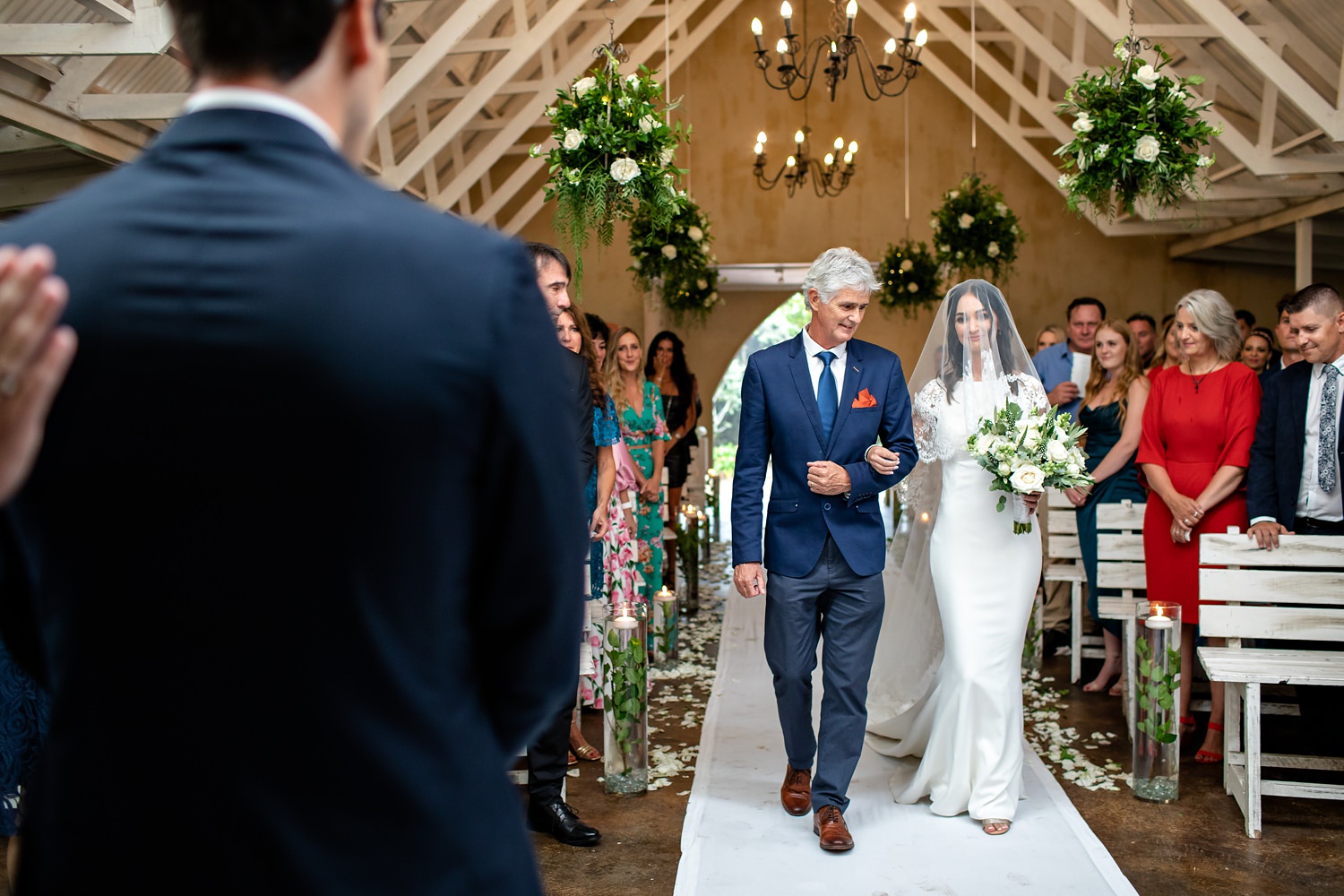 The groom watches as father and daughter walk down the aisle surrounded by smiling guests, flowers and high white ceilings in The Plantation Chapel.