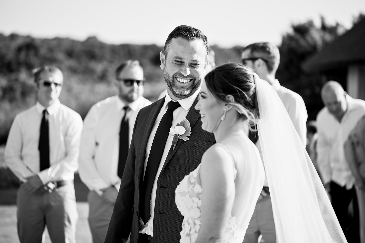 A black and white image of the groom smiling at his bride during their outdoor wedding ceremony