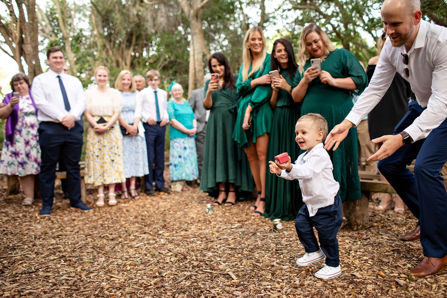 A toddler ringbearer excitedly walks towards the bride and groom with the ring box as smiling wedding guests look on at a forest wedding ceremony in South Africa