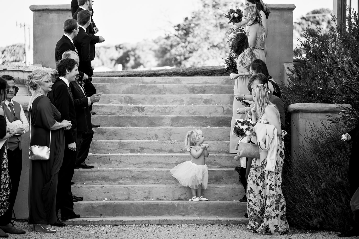 A small flower girl stands on the steps of The Rose Barn as guests line up around her to throw confetti and rose petals on the bridal couple by wedding photographer in South Africa, Niki M Photography