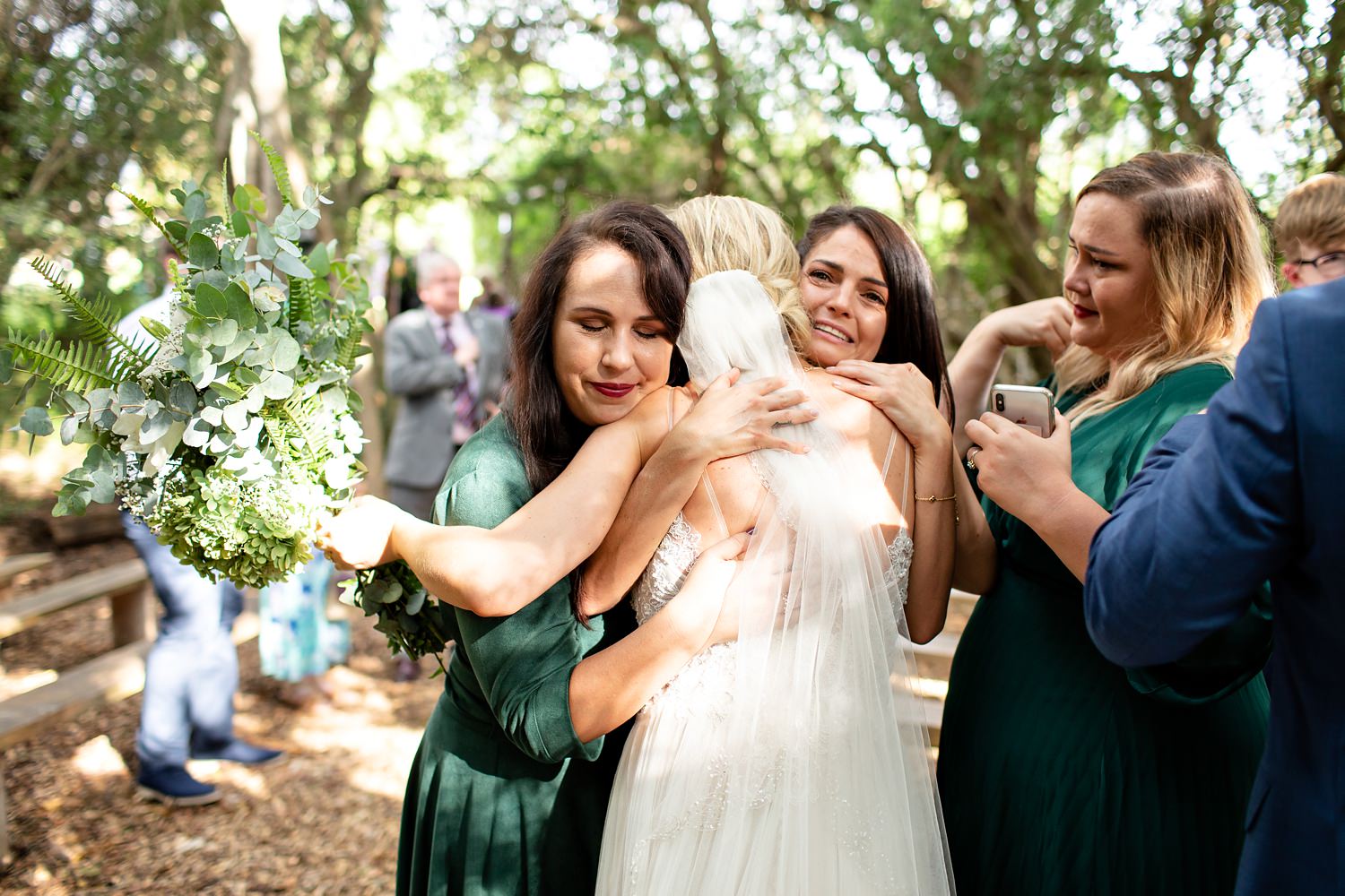 A bride is embraced by her emotional bridemaids dressed in emerald green by wedding photographer in South Africa, Niki M Photography