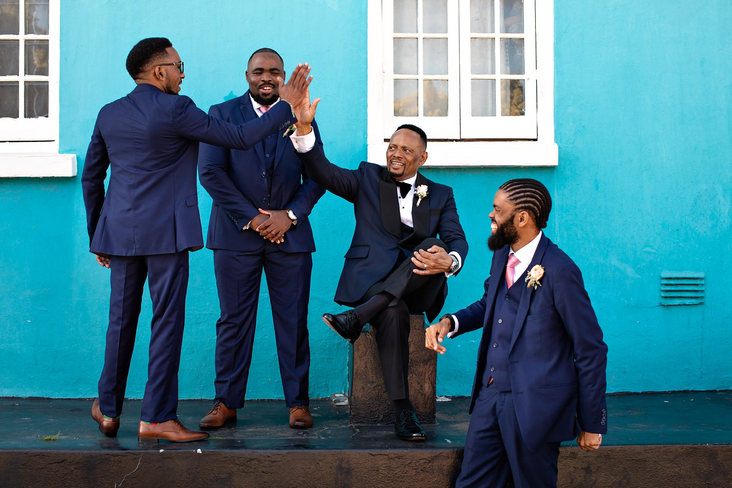 The groom high fives his groomsman in front of a turquoise wall during their bridal party photos by wedding photographer in South Africa, Niki M Photography