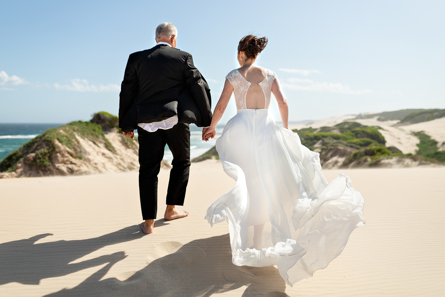 Beach wedding photos. The bride and groom walk over a dune into the wind, with the brides dress blowing out behind her.