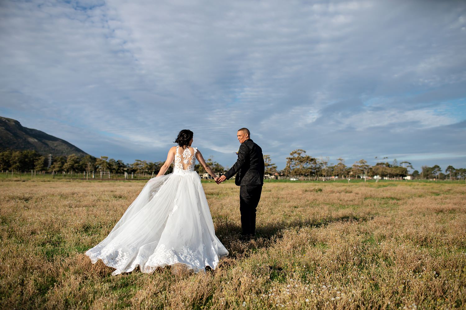 The groom leads the bride into a field, displaying the beautiful train of her wedding dress.