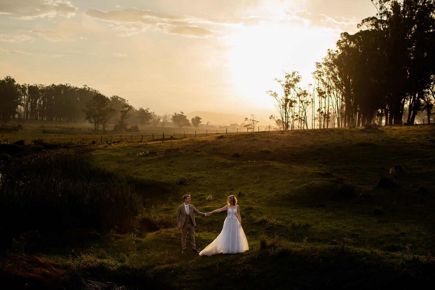 The bride and groom holds hands as the sun sets behind them, illuminating them on the darker landscape