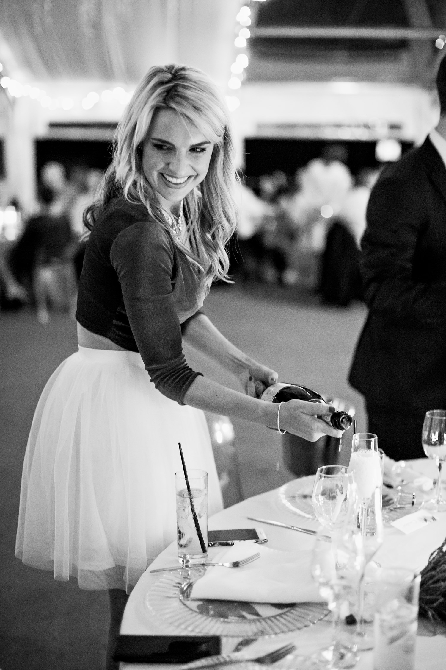 A bridesmaid pours and serves champagne whilst smiling at her husband at Rooftop Garden Bar in Port Elizabeth. Best wedding images by wedding photographer in South Africa, Niki M Photography