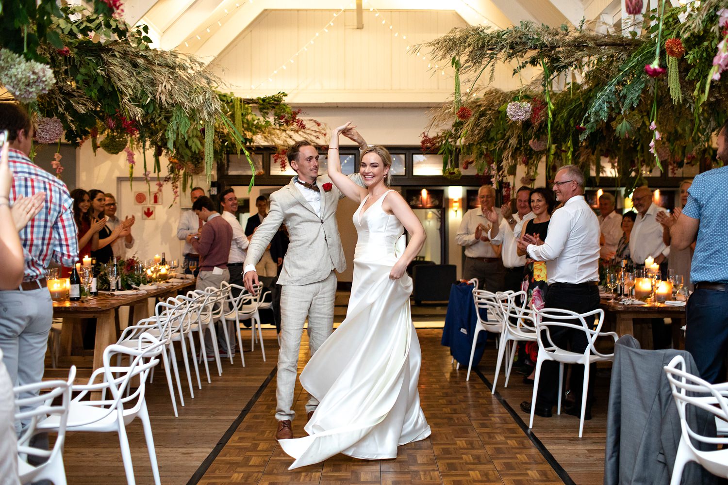 The groom spins the bride during their first dance at The St Francis Links surrounded by clapping guests, a hanging flower installation and contemporary white wedding chairs