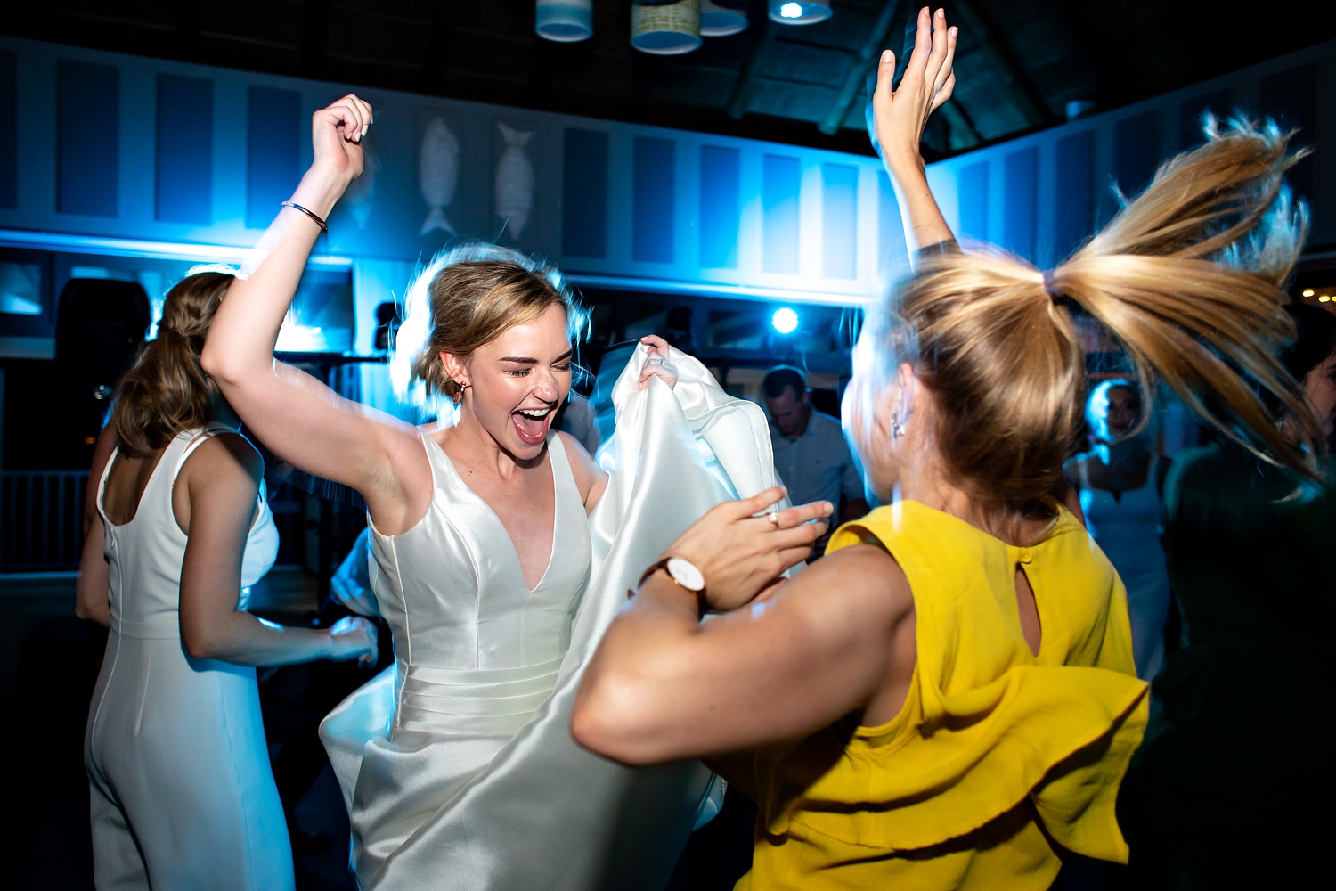 The bride and a guest throw their hands up and jump as the DJ plays amazing music and blue lights flood the wedding dancefloor. Image by wedding photographer in South Africa, Niki M Photography