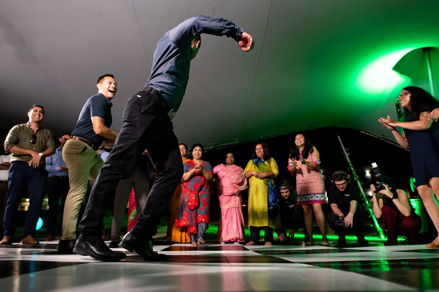 A wedding guest does the moonwalk on a checkered black and white dancefloor as other guests look on and clap 