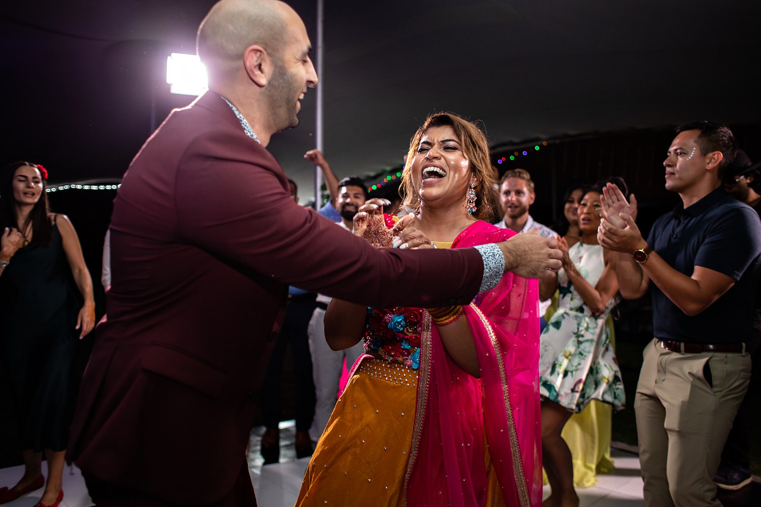 The bride and groom laugh in the middle of the wedding dancefloor dressed in traditional wedding attire and a maroon and gold embellished Sari