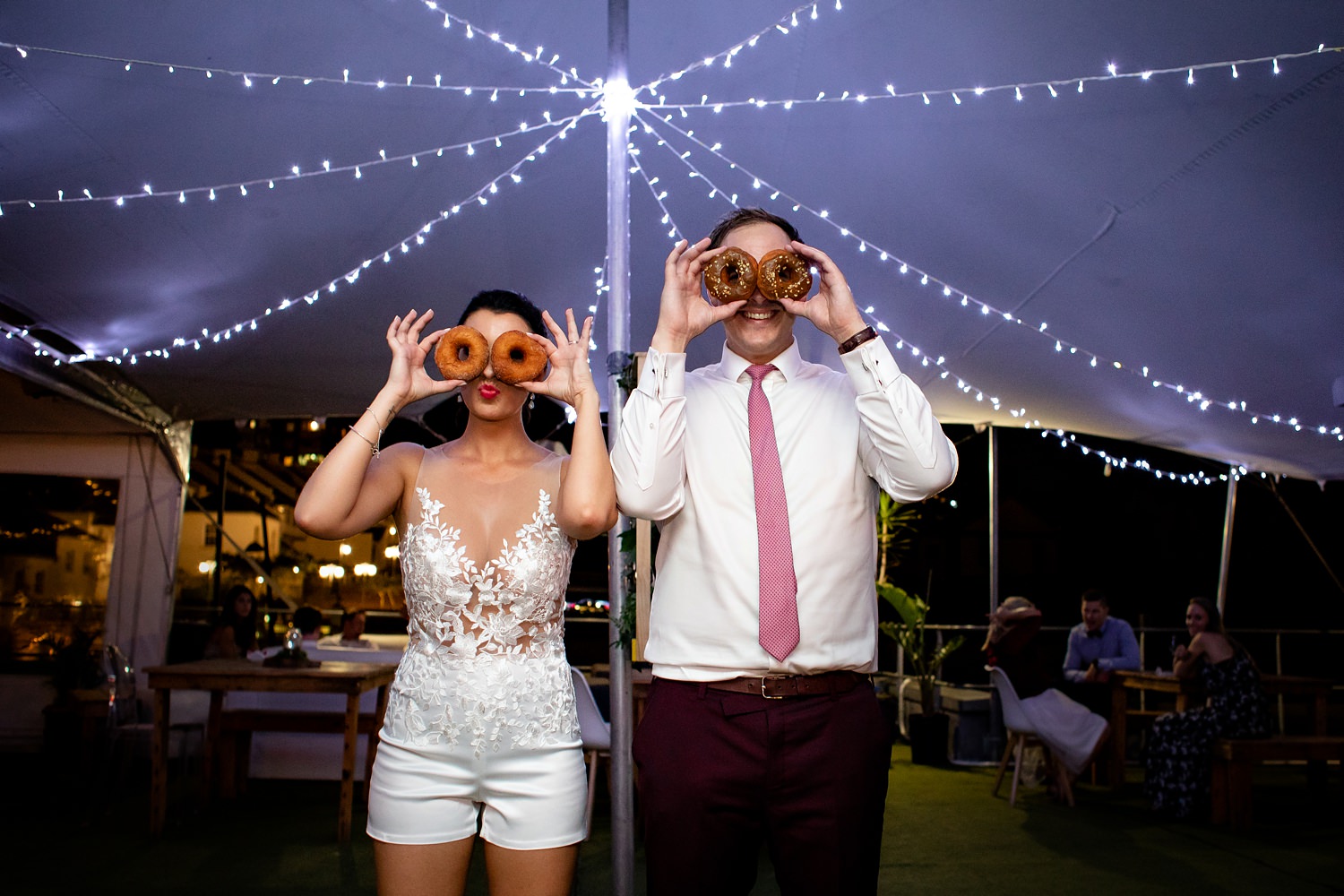 The bride and groom pose at their doughnut dessert bar with doughnuts over their eyes and fairy lights above their heads
