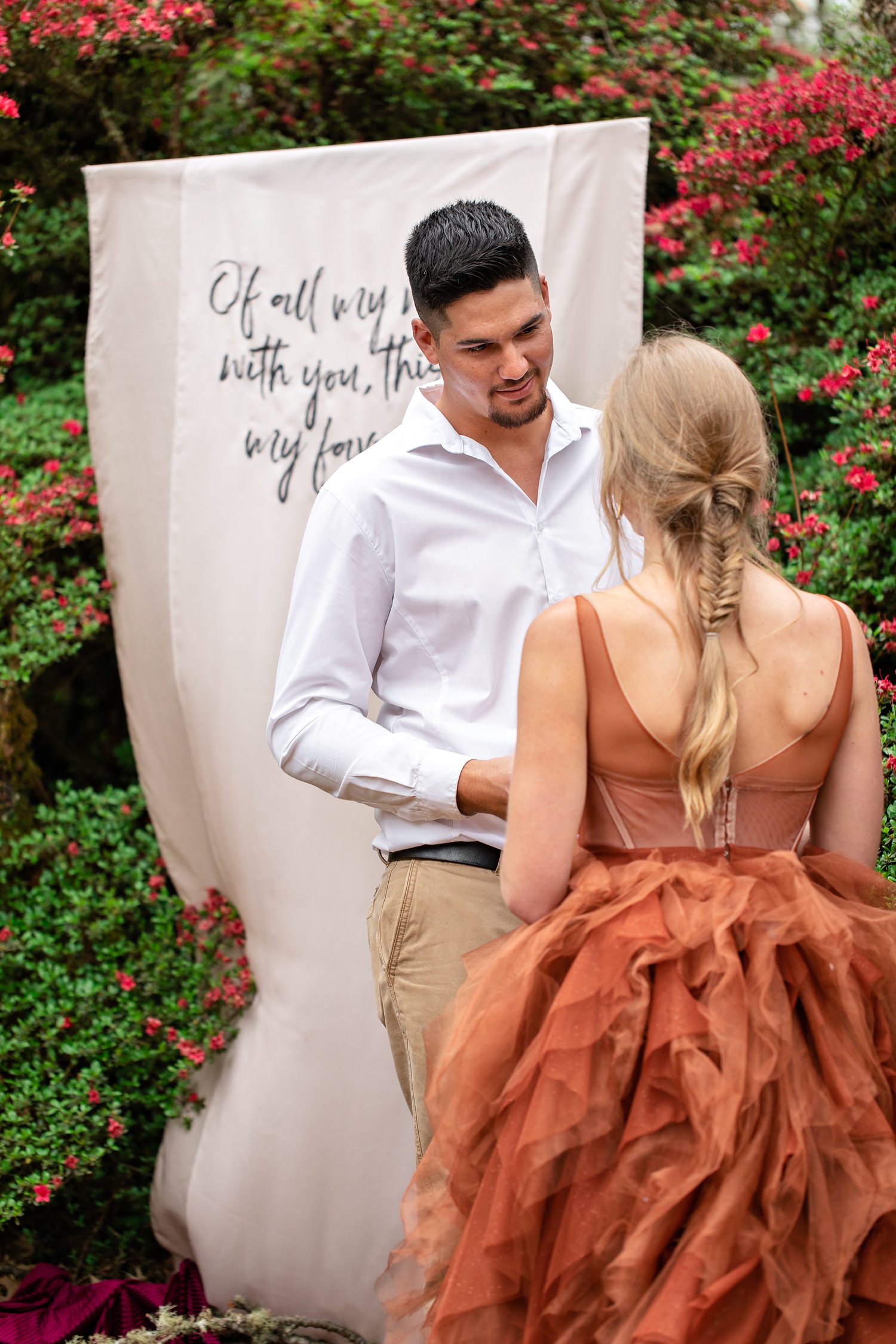 Groom looks at the bride whilst he says his vows with a light pink hanging banner and red flowers behind him.