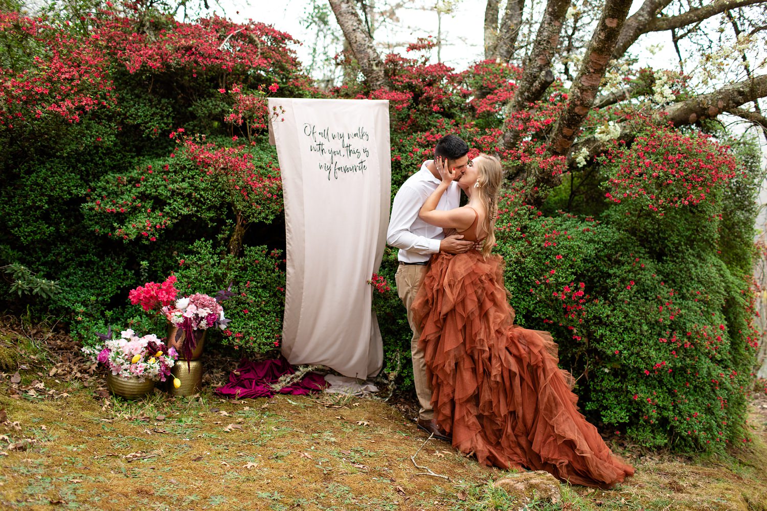 Bride, in a burnt orange wedding dress by Silver Swallow, and Groom kiss at the end of the ceremony in fornt of a DIY banner and azalea flowers in Magoebaskloof, South Africa.