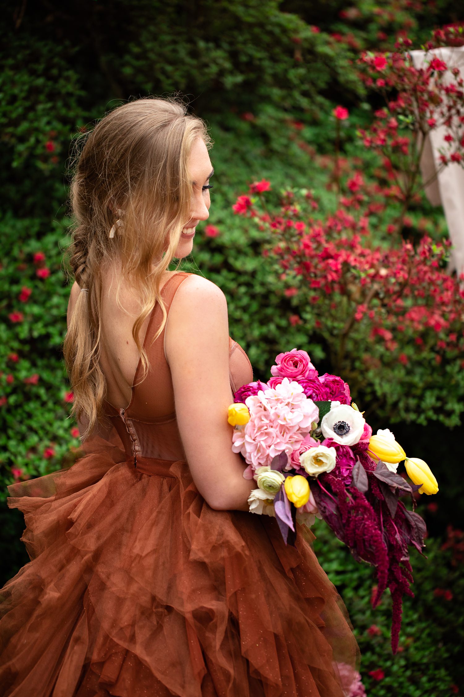 The blonde bride with loose fishtail braid models a colourful bouquet with anemones, country roses, tulips, ranunculus and burgundy amaranth at her Spring elopement.