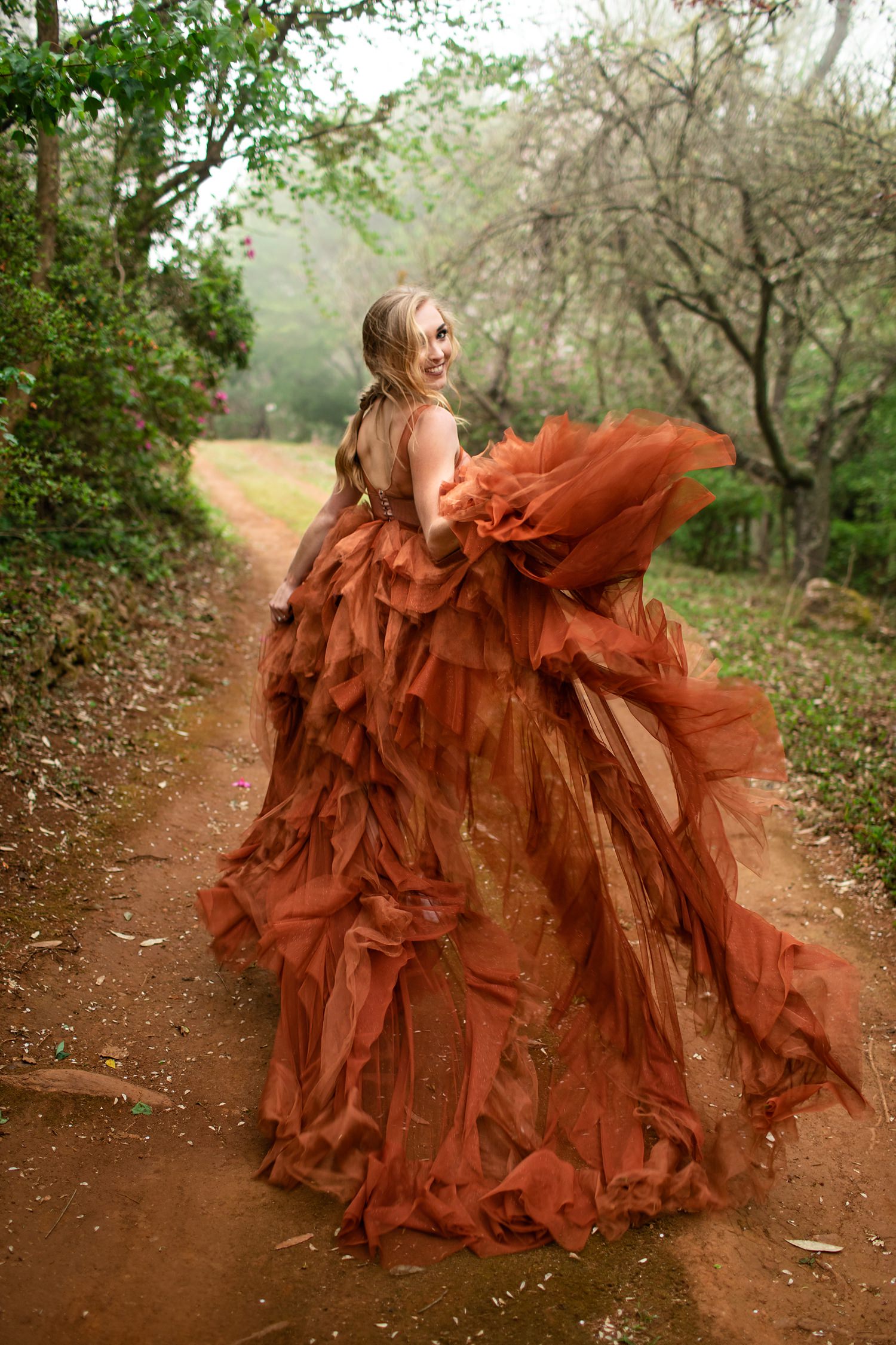 The blonde bride with loose fishtail braid models a burnt orange wedding dress as she runs down a tree-lined dirt road at her elopement in Magoebaskloof