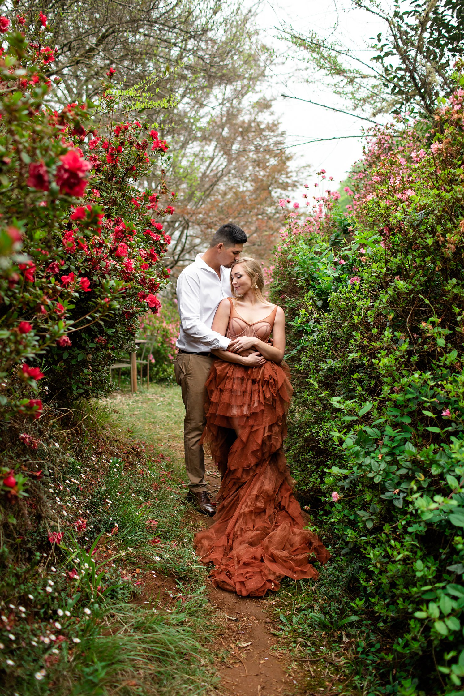 The Bride and Groom pose in between hedges of Azaleas for elopement wedding photographer Niki M in Magoebaskloof. She wears a beautiful alternative flowing wedding gown in burnt orange 