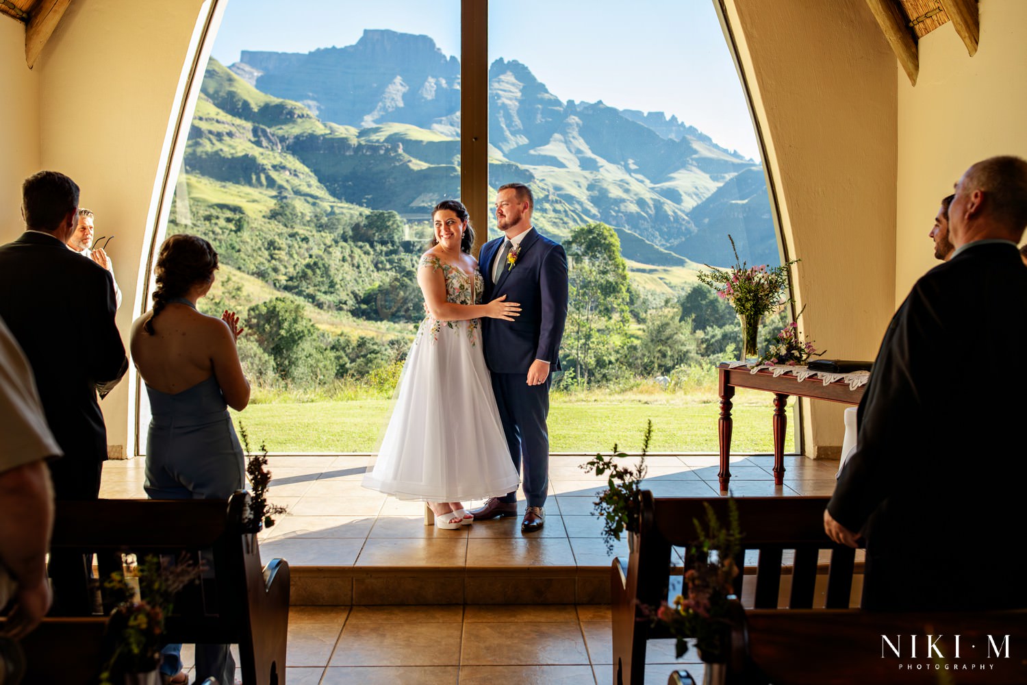 Bride and groom standing in front of the iconic Champagne Castle Hotel chapel window, framed by the sweeping Drakensberg mountain view behind them, celebrating their intimate Drakensberg Micro-Wedding ceremony.