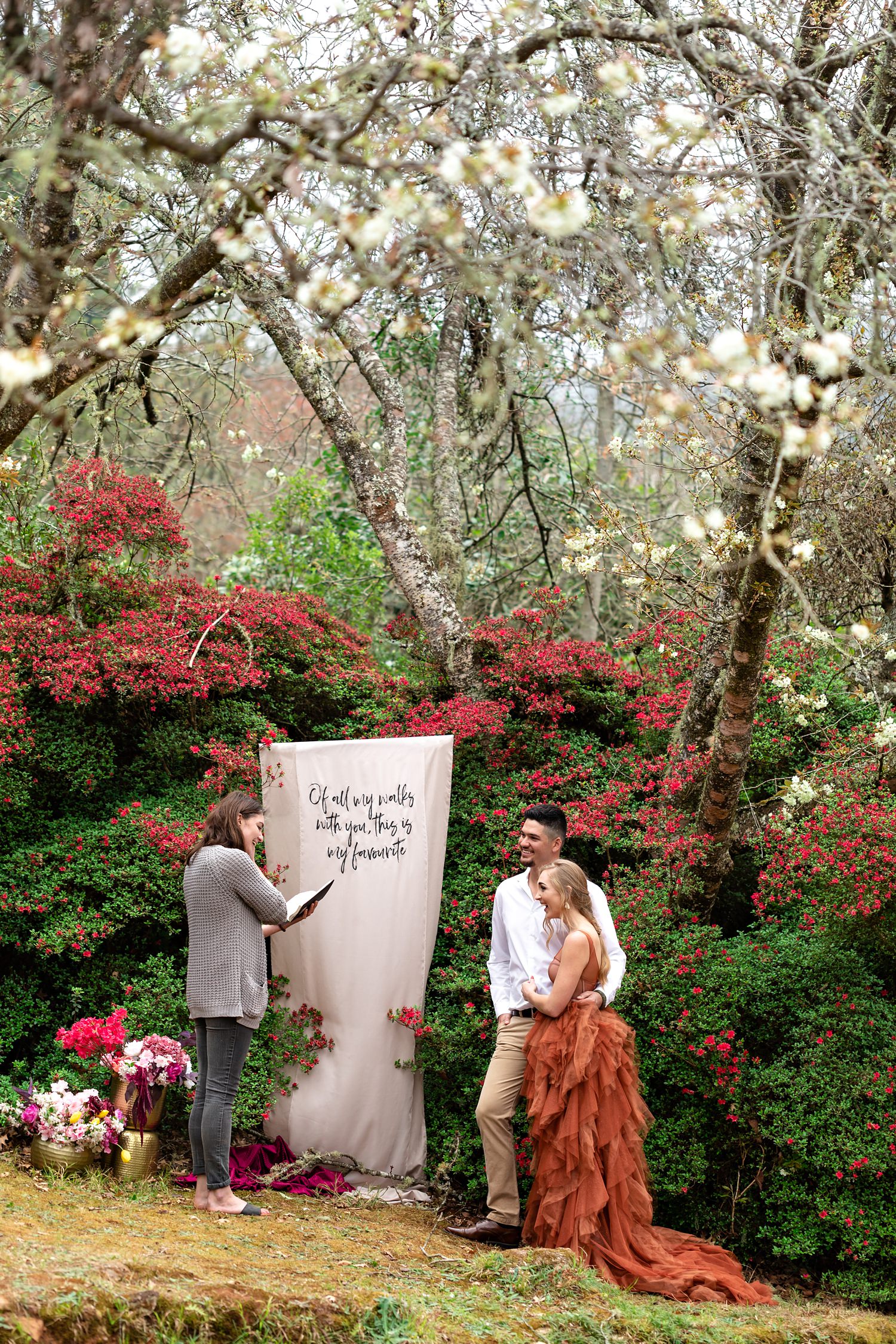 Bride, in a burnt orange wedding dress by Silver Swallow, and Groom kiss at the end of the ceremony in fornt of a DIY banner and azalea flowers at their elopement in Magoebaskloof, South Africa.