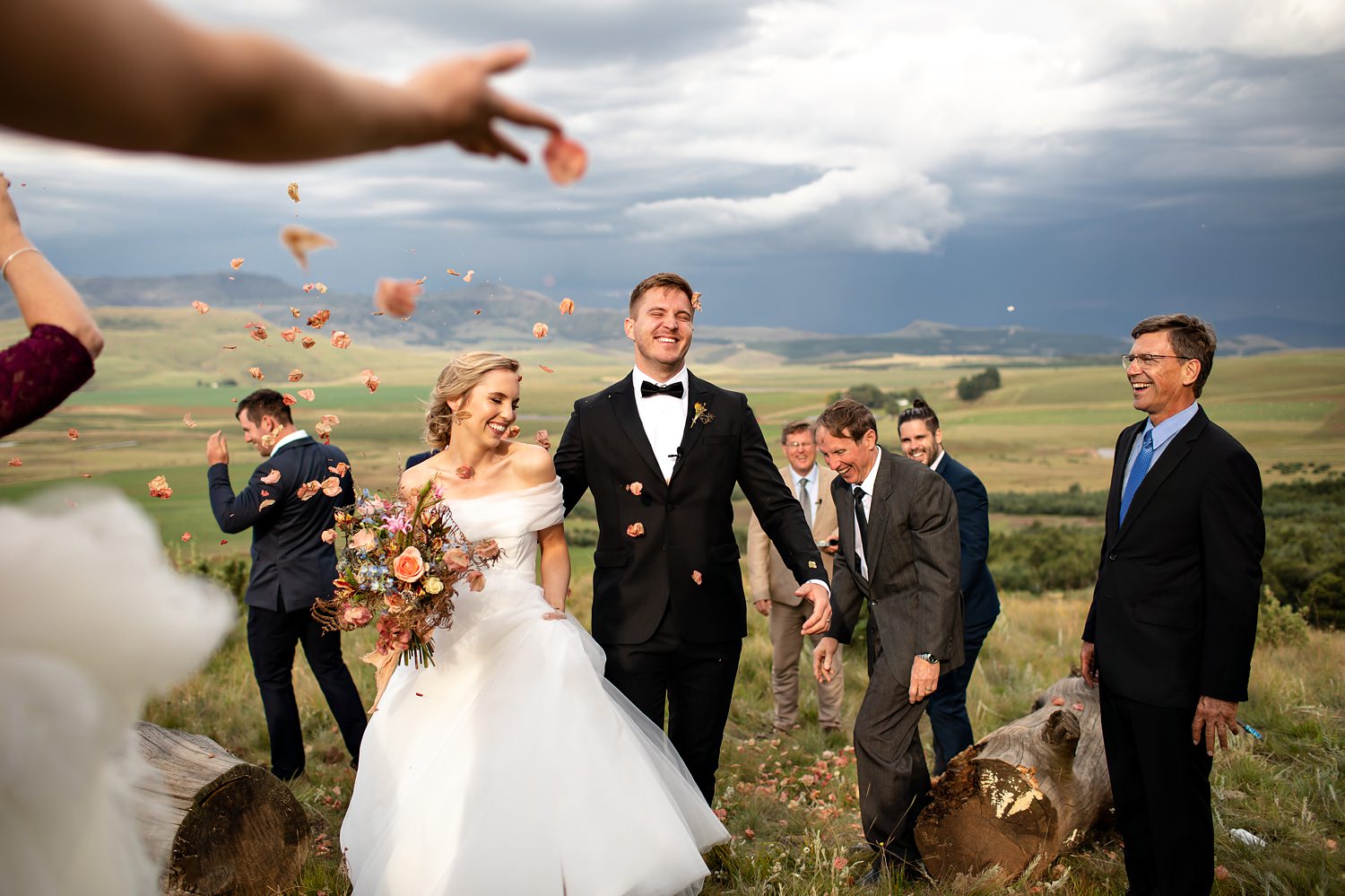 Guests toss dry bougainvillea and hydrangea flowers as confetti at the end of a Drakensberg wedding ceremony, with the mountains behind the couple.