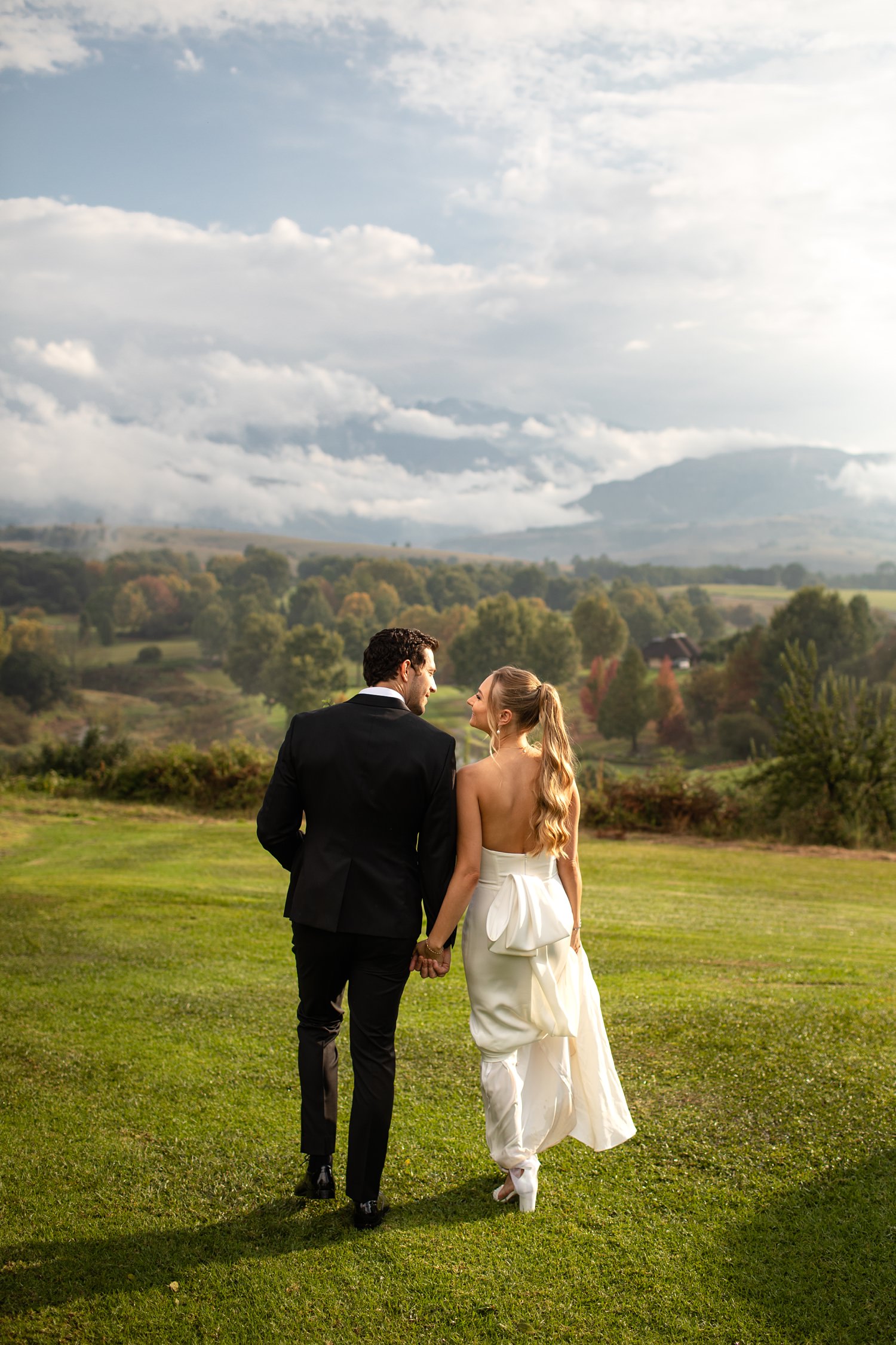Beautiful Autumn scenery with views over the Champagne Sports Golf Course and Central Drakensberg mountain range as the bride and groom walk away from the camera at their wedding.