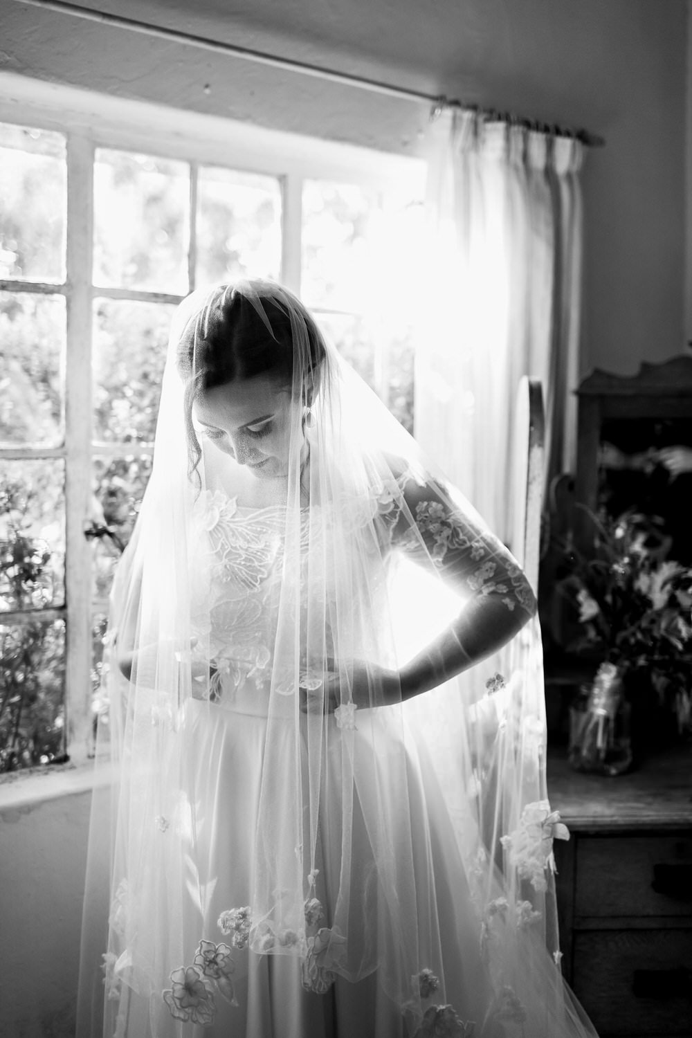 A beautiful black and white image of a bride in a veil, standing in front of a cottage window, as she makes final adjustments to her gown before her Elgin wedding