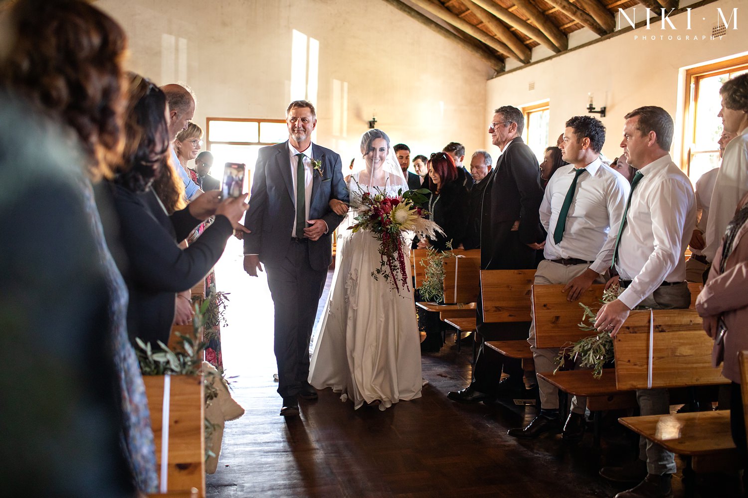 Bride walks down the aisle accompanied by her father in a small chapel on the Paul Cluver wine estate for her Elgin wedding