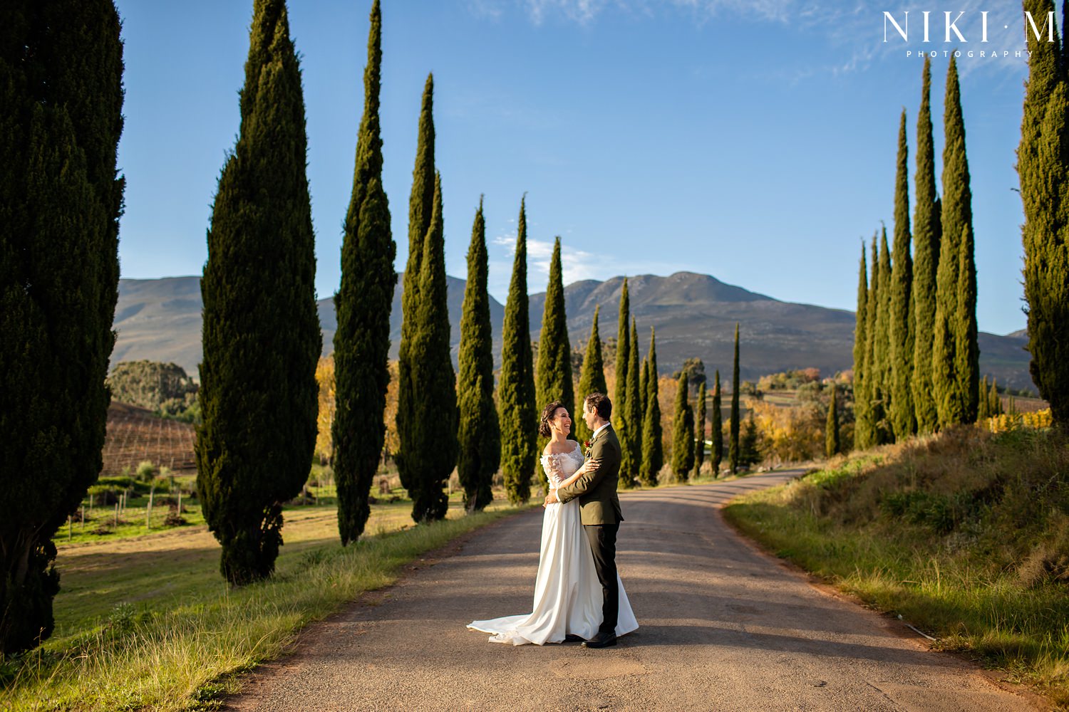 Cypress trees and the bridal couple after their ceremony at Paul Cluver Wine's chapel for their Elgin wedding. The bride wears a beautiful dress by Fender Bridal Design.