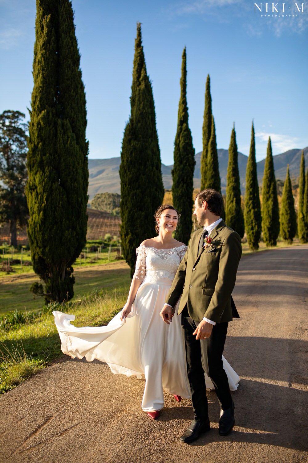 Cypress trees and the bridal couple after their ceremony at Paul Cluver Wine's chapel for their Elgin wedding. The bride wears a beautiful dress by Fender Bridal Design and red shoes.