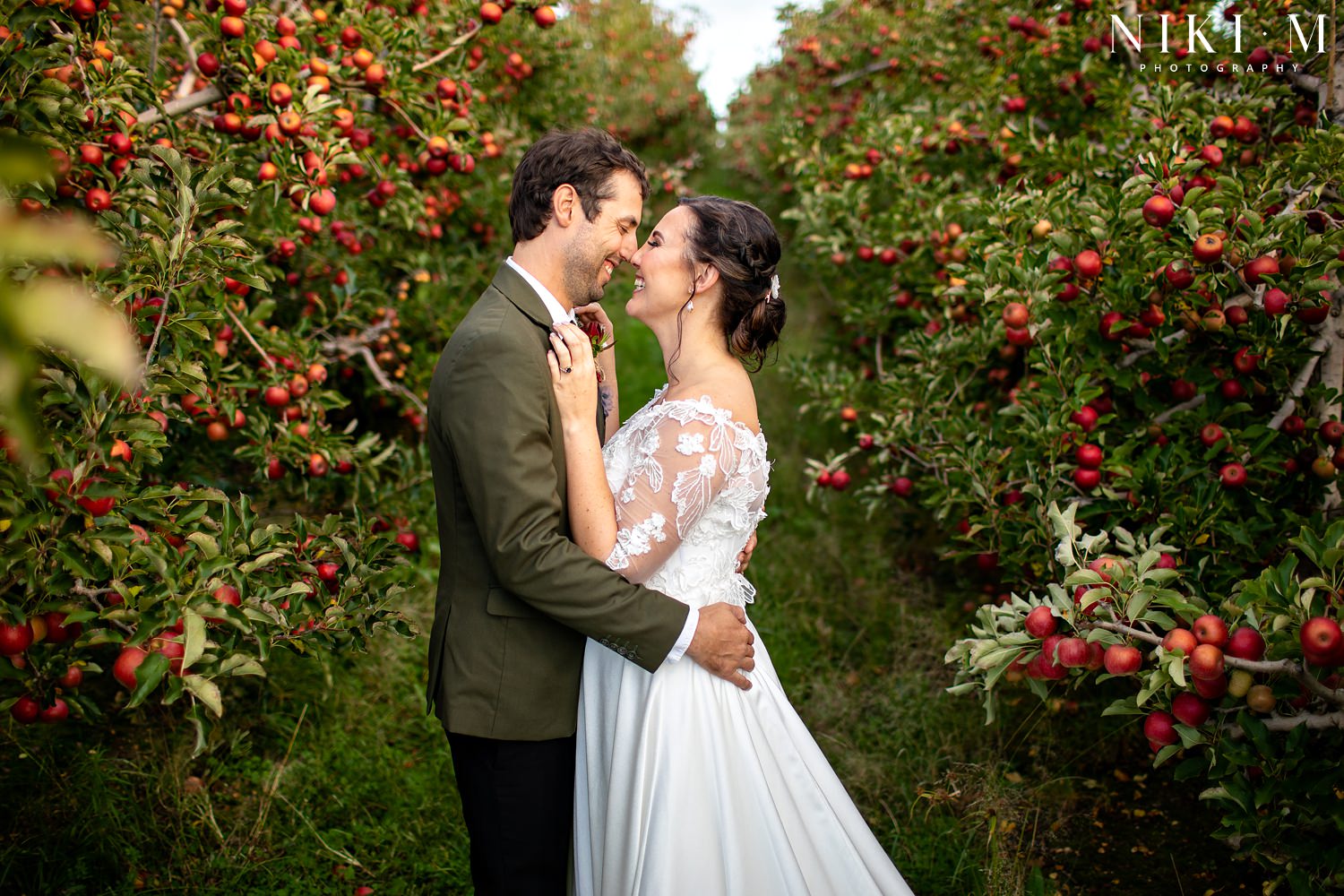 The bride and groom touch noses and laugh whilst surrounded by red apple trees in an apple orchard for their Elgin wedding at Cherry Glamping