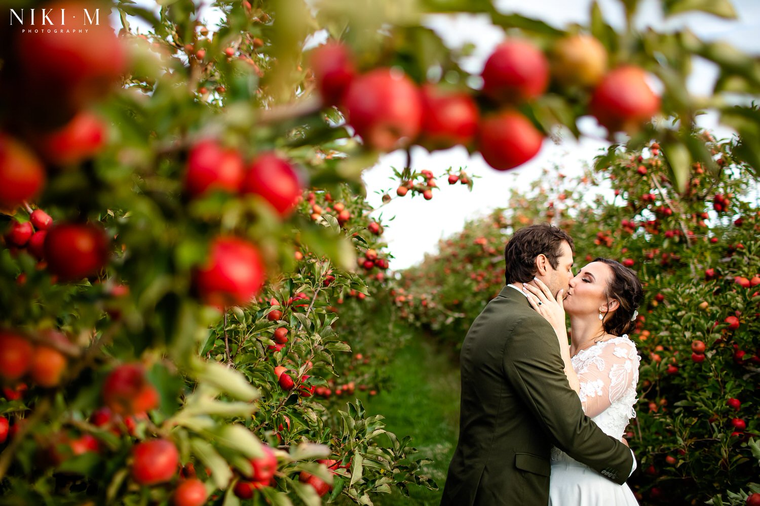 A bride and groom kiss whilst framed by red apple trees in an apple orchard for their Elgin wedding at Cherry Glamping