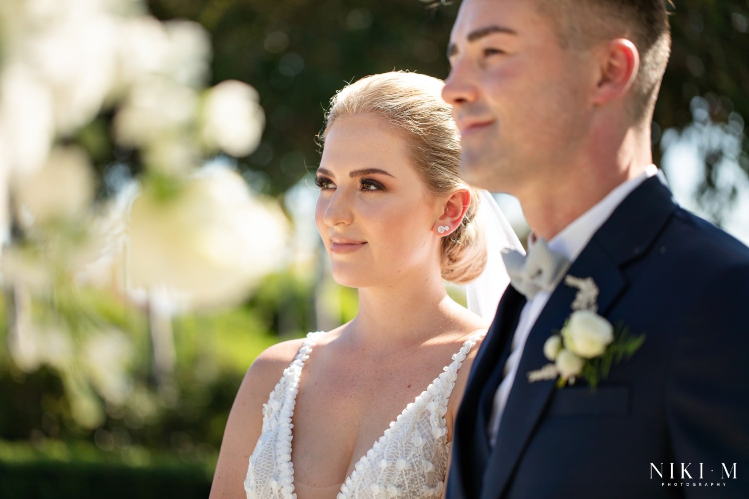 The bride wears copper and brown toned make-up and a low slung neat bun for her Cavalli Estate wedding in the sunken garden with white rose floral arch
