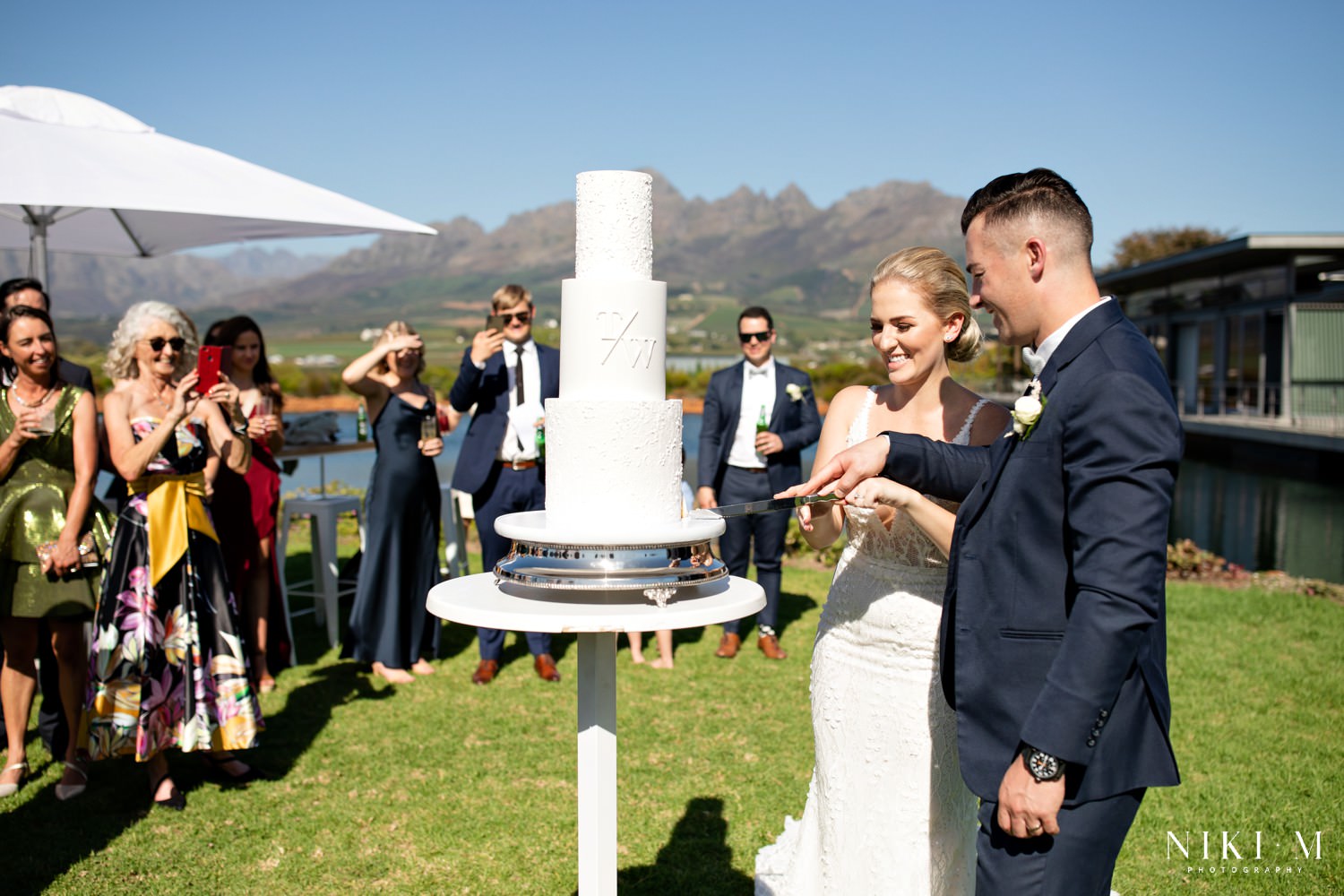 Cavalli Estate wedding cake cutting during canapes. Three tier classic white wedding cake with minimalist design and monogram
