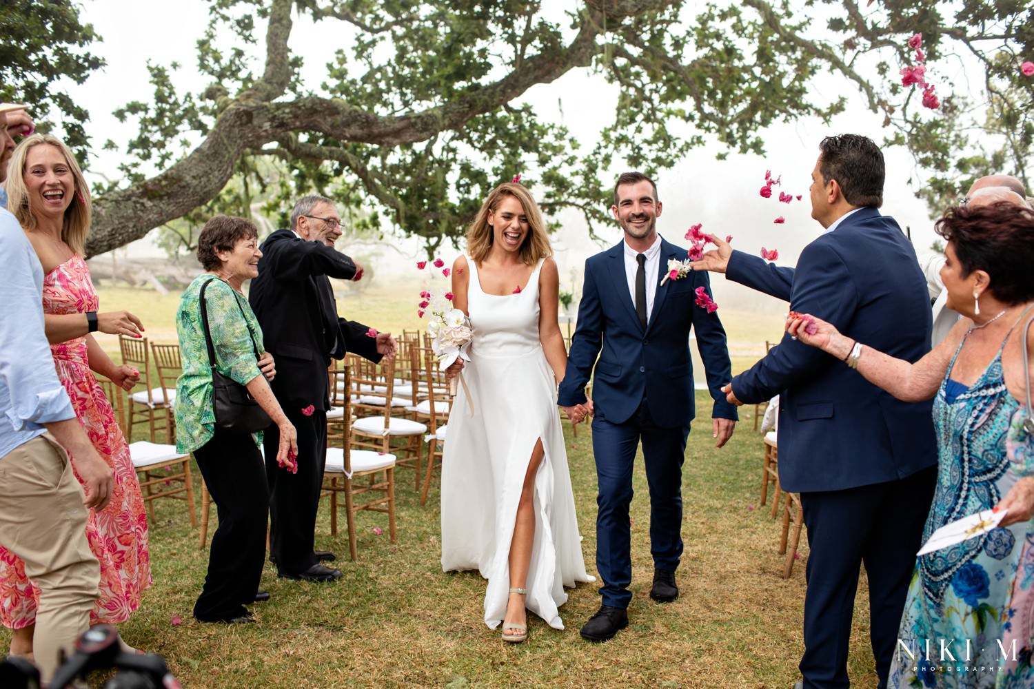 The bride and groom walk through their cheering guests and are showered with pink bougainvillea flowers after their wedding ceremony.