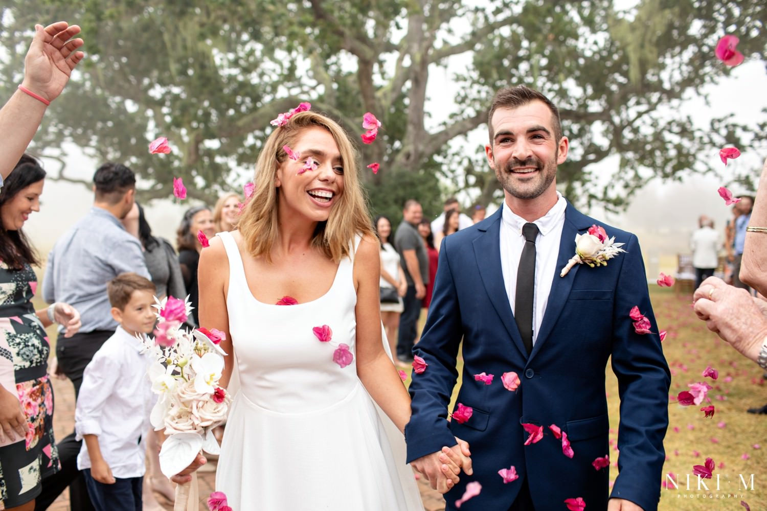 The bride and groom walk through their cheering guests and are showered with pink bougainvillea flowers after their wedding ceremony.