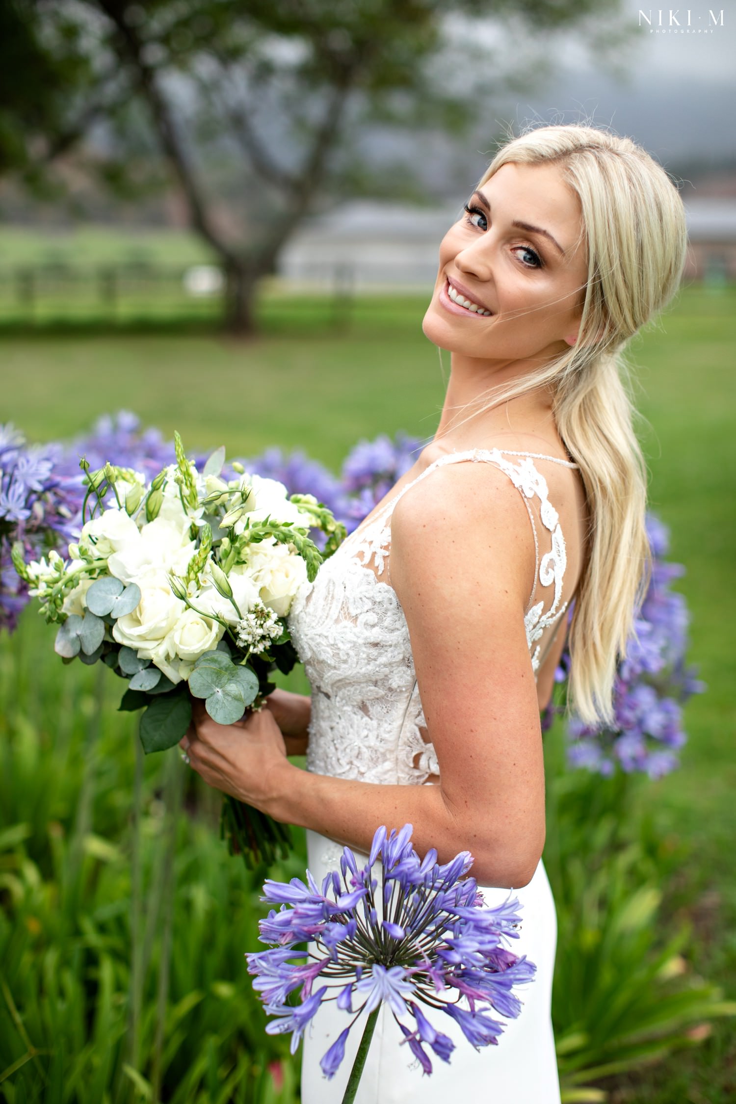 Bridal portrait amongst the purple agapanthus, showing wedding dress with lace illusion neckline.