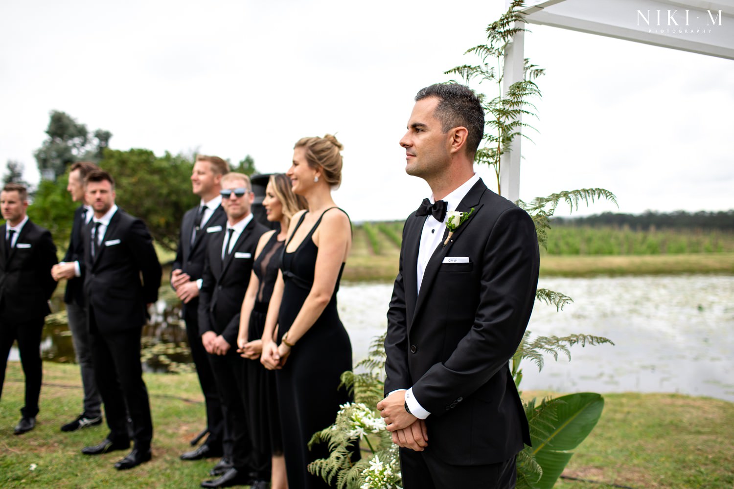 Groom wears formal black tie for his outdoor wedding, surrounded by his bridal party also dressed in black.