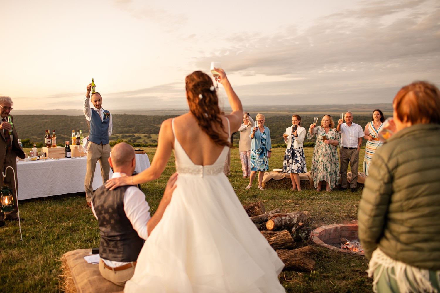 Toasts at sunset. The bridal party gather around a fire pit in the South African bush at a wedding. Image by safari photographer Niki M