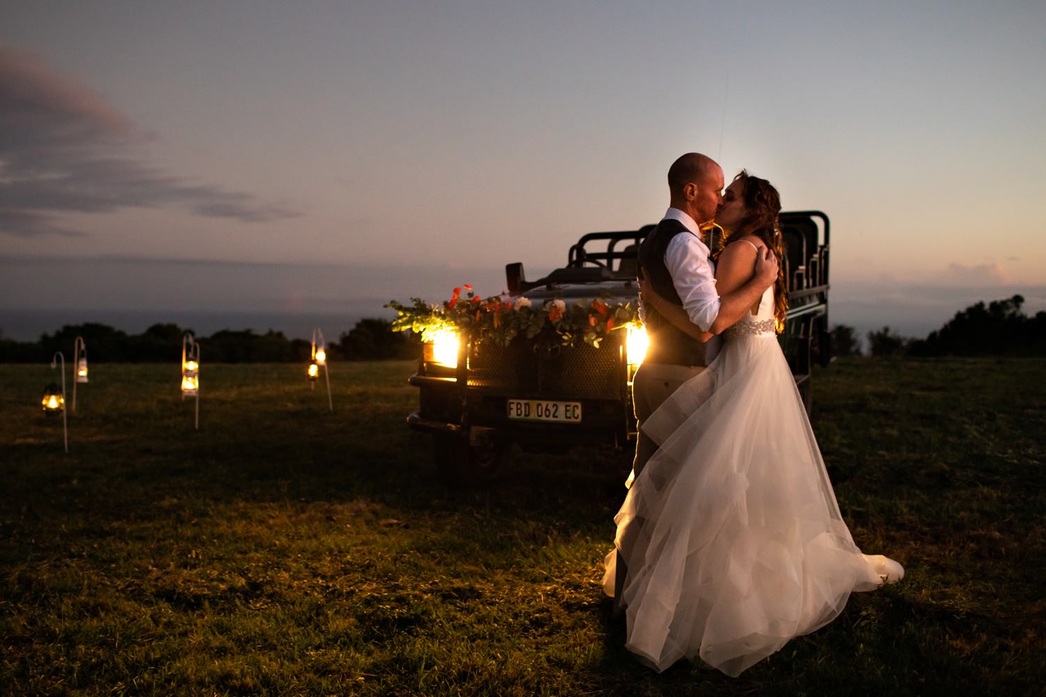 A bride and groom embrace in front of a game vehicle, lit up by the lights of the vehicle and candle lanterns, at their bush wedding. An image by safari photographer Niki M