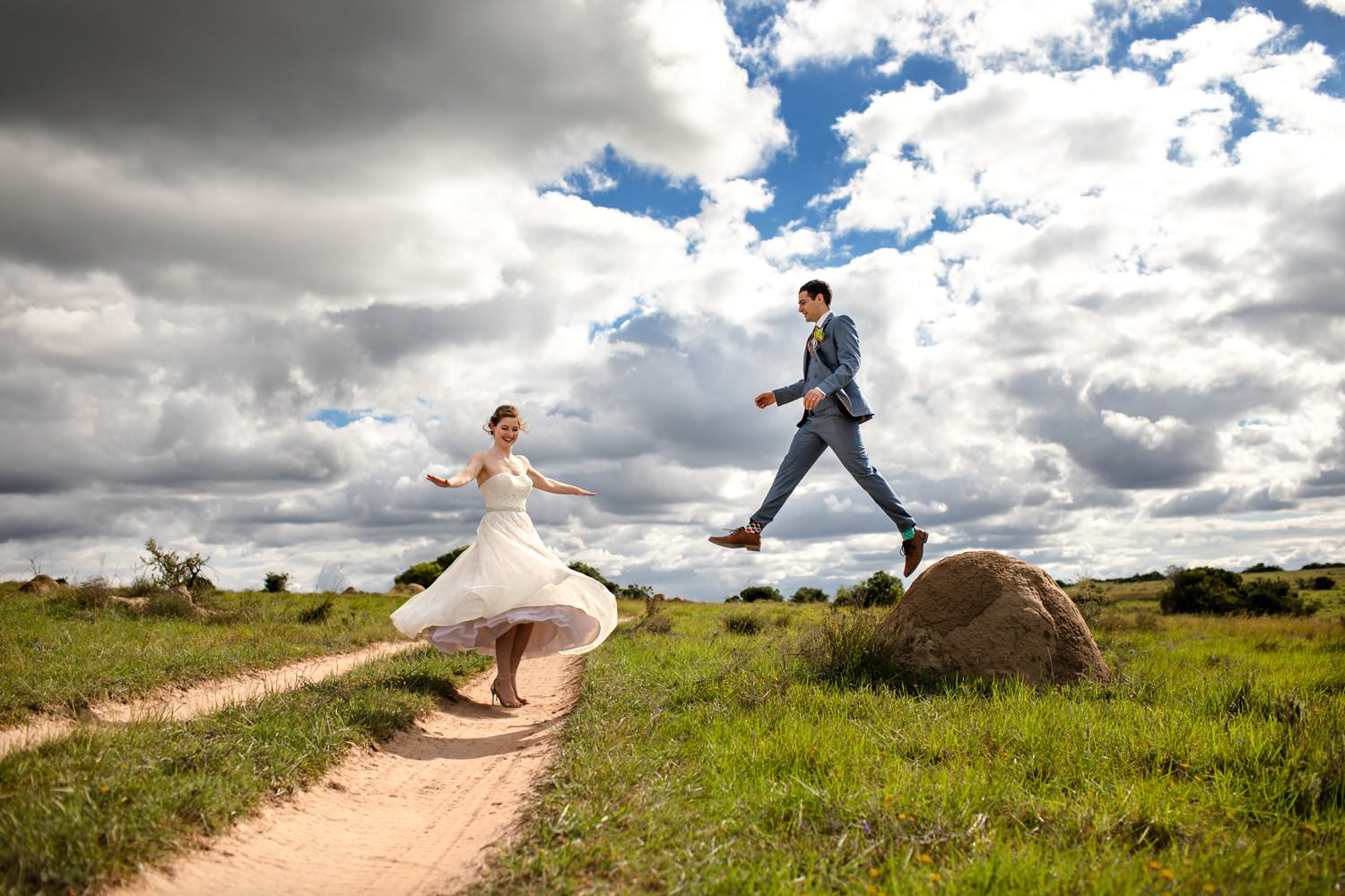 Safari wedding photographer Niki M captures a laughing groom jumping off an anthill whilst the bride spins in her mid-length wedding dress at their destination bush wedding in South Africa