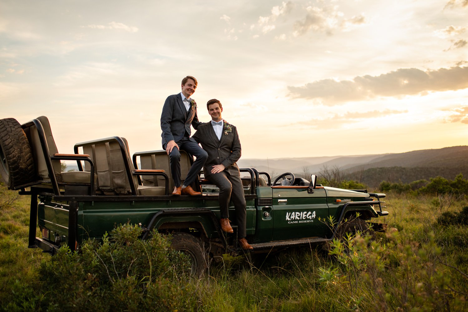 Image of two grooms posing on a Kariega Game Lodge vehicle with the sunset behind them at their Safari Elopement by Safari Photographer Niki M