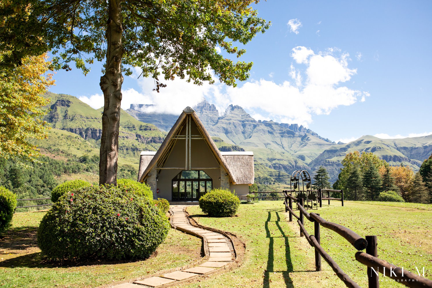 The thatched glass chapel at Champagne Castle Hotel surrounded by lush green lawns and the dramatic Drakensberg mountains on a clear autumn morning — a breathtaking setting for a Drakensberg micro-wedding ceremony.
