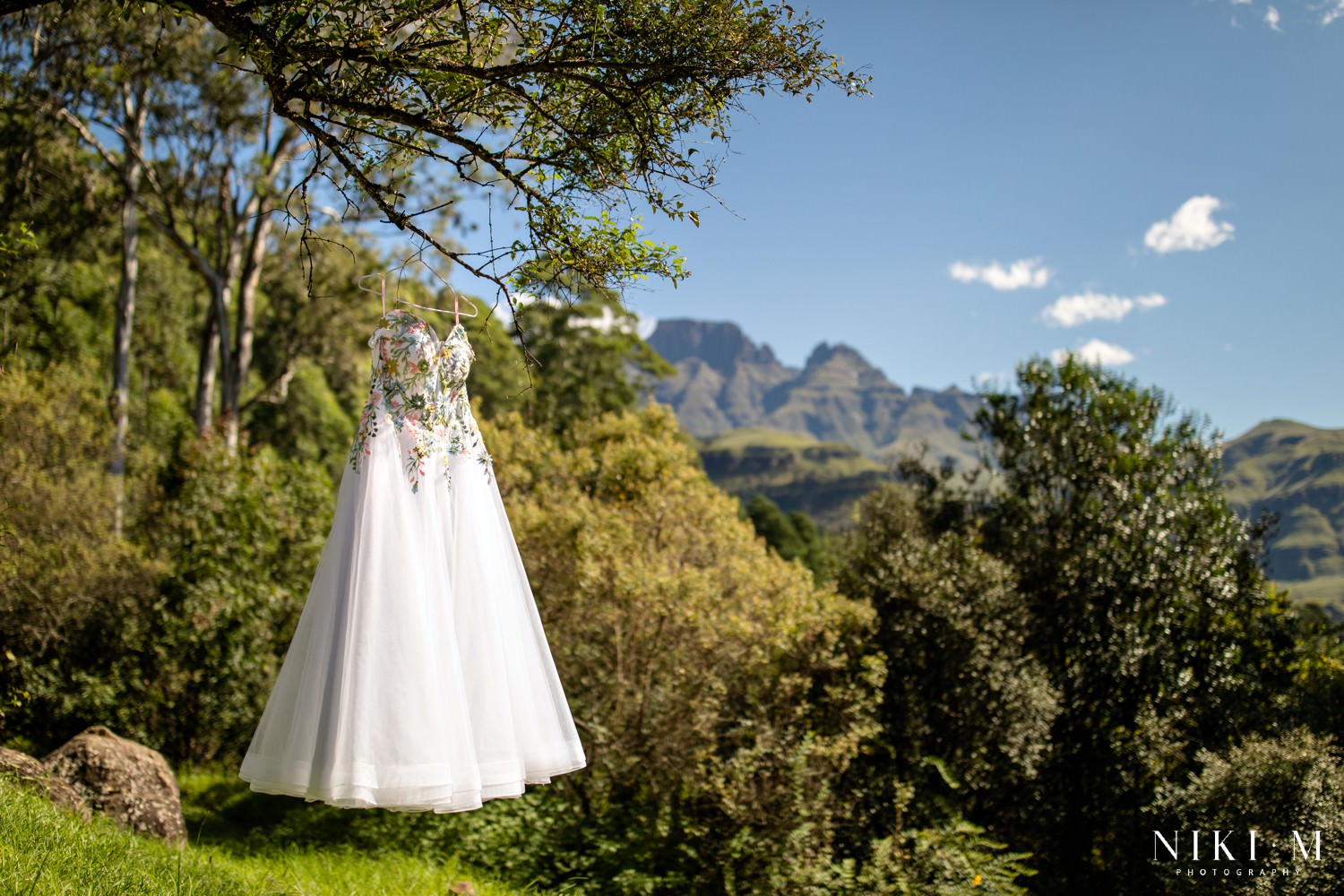 An embroidered floral wedding dress hanging from a tree branch with the Drakensberg peaks in the background — a whimsical detail shot capturing the essence of a romantic Drakensberg micro-wedding.